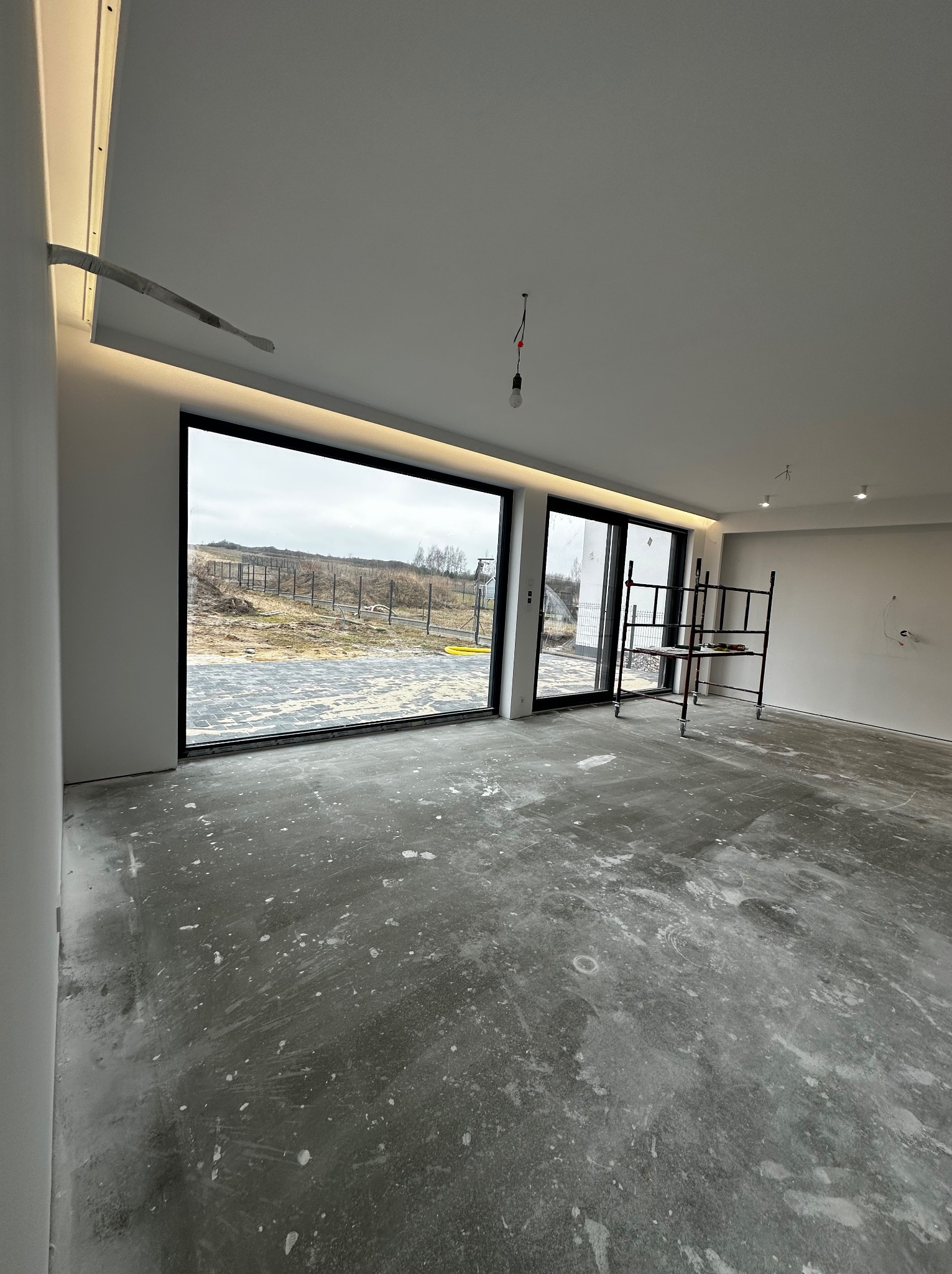 Surowy stan wnętrza z dużym oknem i rusztowaniem. Podłoga z widocznymi śladami prac, przygotowana do dalszych etapów wykończenia. Minimalistyczne oświetlenie sufitowe.