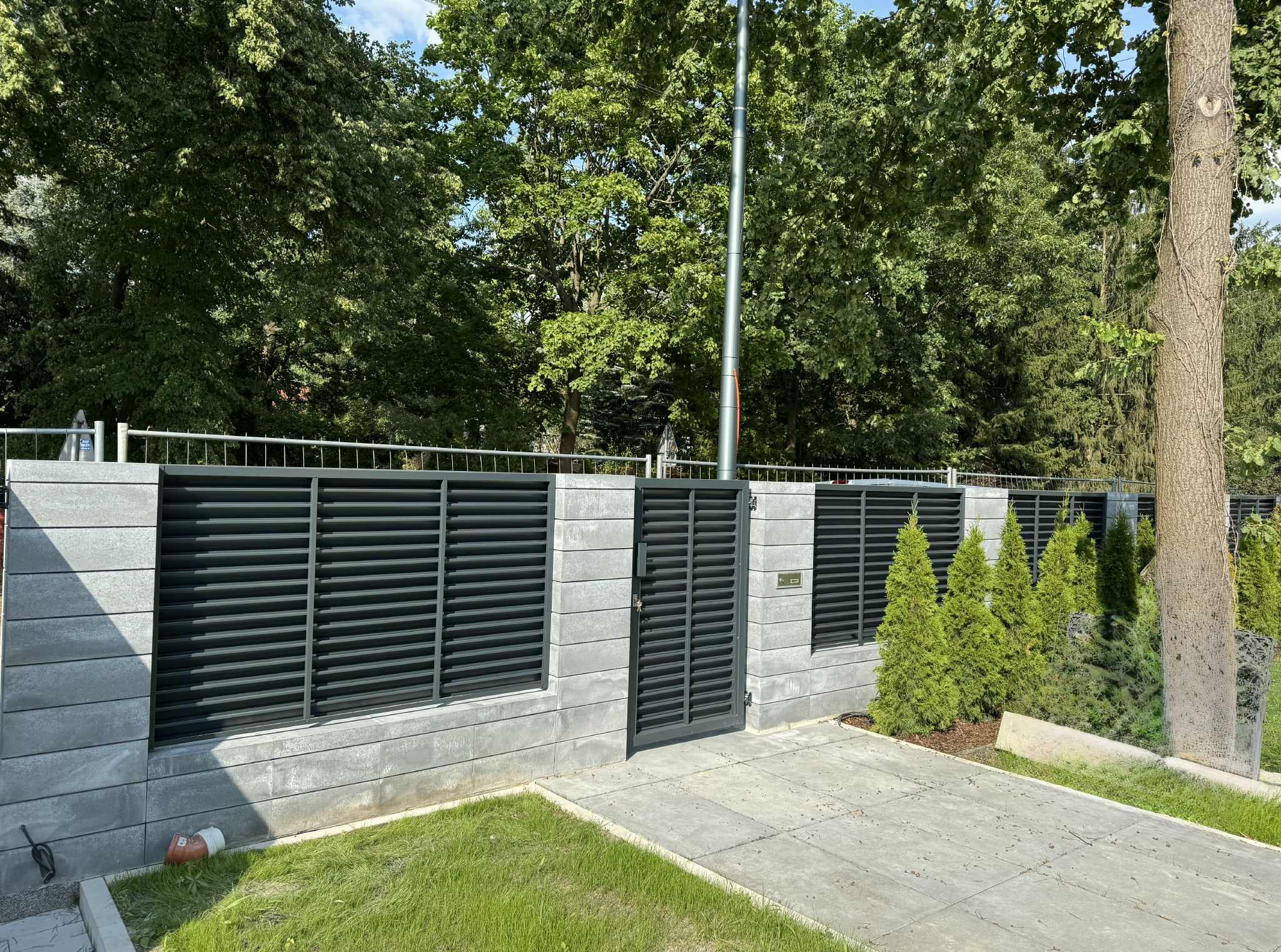 Nowoczesne ogrodzenie z szarych bloczków betonowych i czarnych, poziomych paneli metalowych. Brama wejściowa w kolorze paneli. Tuje posadzone wzdłuż ogrodzenia.