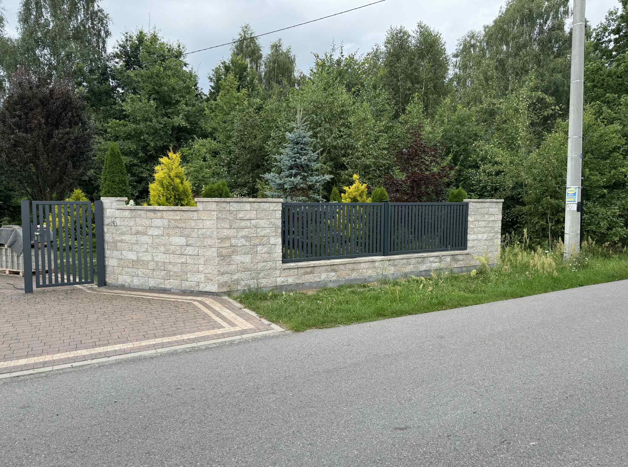 Nowoczesne ogrodzenie z metalowymi przęsłami i elementami z jasnego kamienia łupanego, z bramą w tle, otoczone zielenią i drzewami. Widok z poziomu ulicy.