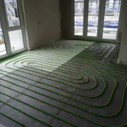 Zielone rury ogrzewania podłogowego ułożone na czarnej folii w surowym wnętrzu z oknami, przez które widać rusztowania budowlane.