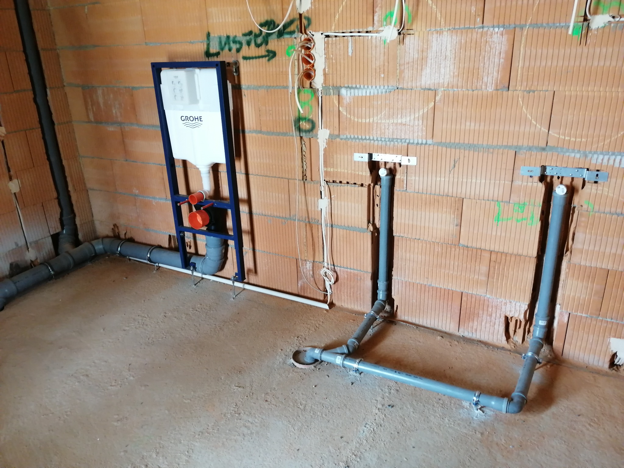 Instalacja podtynkowa WC Grohe w surowym wnętrzu. Widoczne szare rury kanalizacyjne i instalacja wodna na tle ceglanej ściany w trakcie montażu.