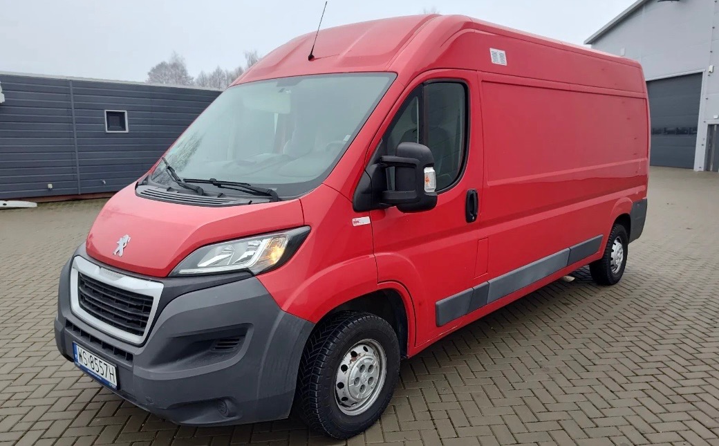 Czerwony Peugeot Boxer z wysokim dachem na brukowanym placu; widoczne detale jak tablica rejestracyjna i logo. W tle szara hala i kontener biurowy.