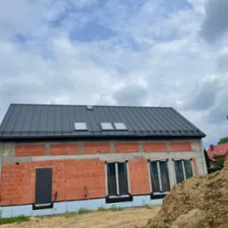 Surowy, ceglany budynek z szarą blachą, trzema oknami dachowymi i czarnymi oknami. Widoczne materiały budowlane i ziemia. Etap budowy domu jednorodzinnego.