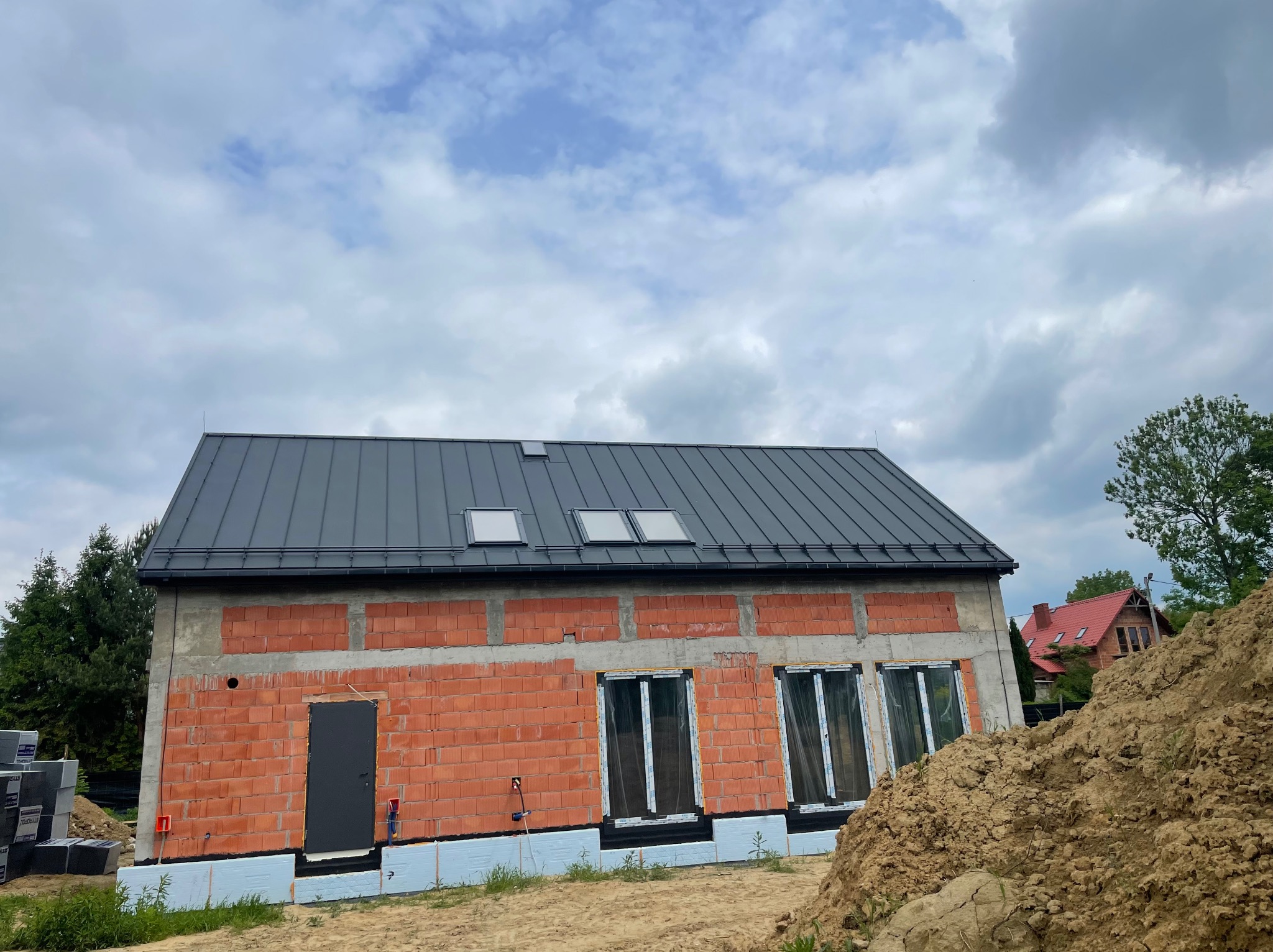 Surowy, ceglany budynek z szarą blachą, trzema oknami dachowymi i czarnymi oknami. Widoczne materiały budowlane i ziemia. Etap budowy domu jednorodzinnego.