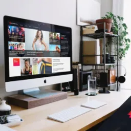 Biurko z komputerem iMac wyświetlającym stronę mysportlife.pl z artykułami o zdrowym stylu życia i ćwiczeniach, obok szklanka wody, słuchawki i organizer z przyborami biurowymi.