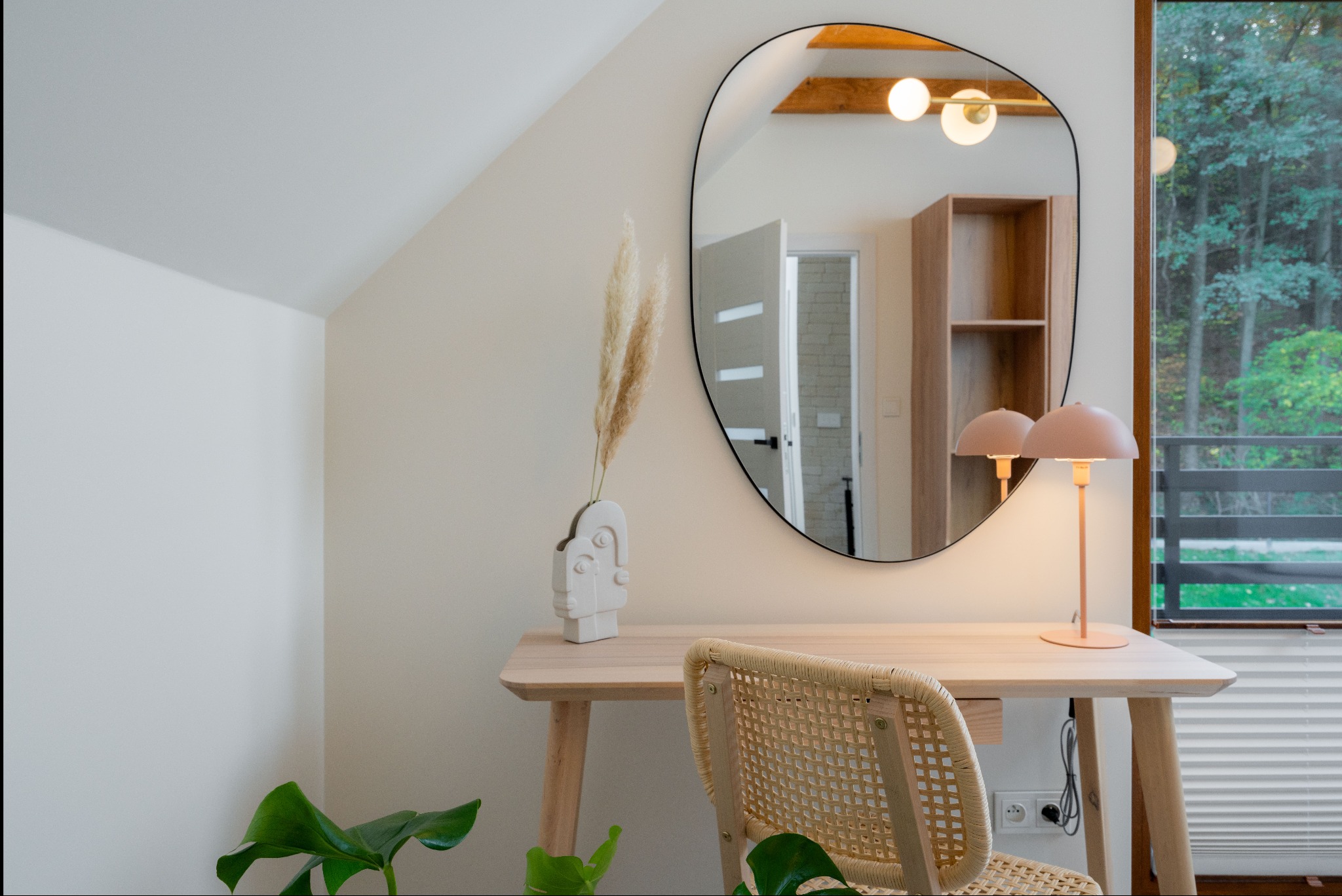 Jasne wnętrze pokoju z biurkiem, wiklinowym krzesłem, lampką i lustrem, w którym odbija się widok na las. Neutralna kolorystyka, naturalne materiały, minimalistyczny styl.