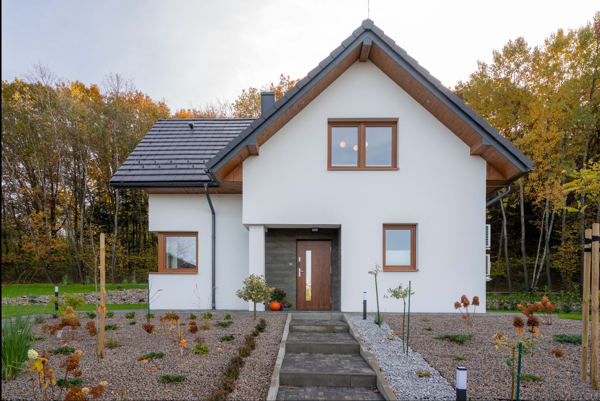 Biały dom z brązowymi oknami, ciemnym dachem i brukowanym chodnikiem. Jesienne drzewa w tle. Dynia przed wejściem dodaje akcentu.