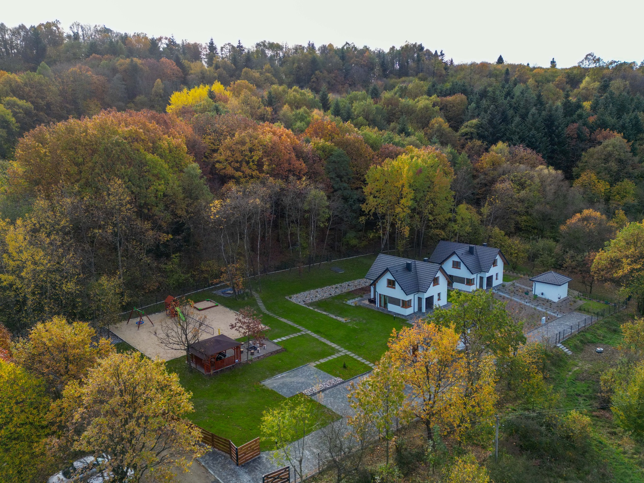 Jesienny widok z lotu ptaka na osiedle domów jednorodzinnych z placem zabaw, otoczone lasem w barwach jesieni. Uwagę przykuwa symetryczny układ ścieżek i trawników.