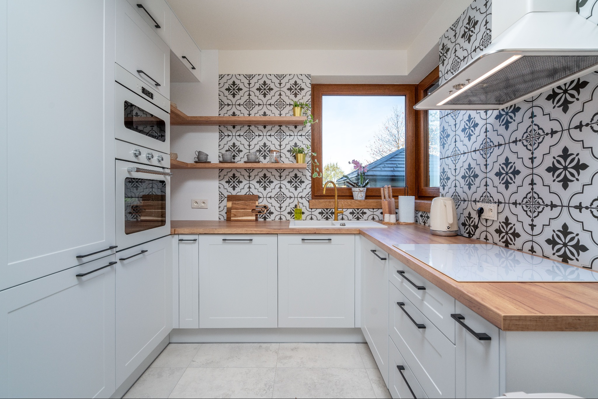 Biała kuchnia z drewnianymi blatami, wzorzystymi płytkami i oknem z widokiem na zieleń. Nowoczesny design z elementami retro, złota bateria i minimalistyczne dodatki.