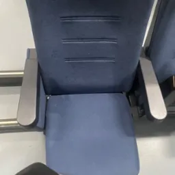 Granatowe, pluszowe krzesło z podłokietnikami. Widok z góry na czyste siedzisko. Fragment ubrania w lewym dolnym rogu kadru. Krzesło w przestrzeni publicznej.