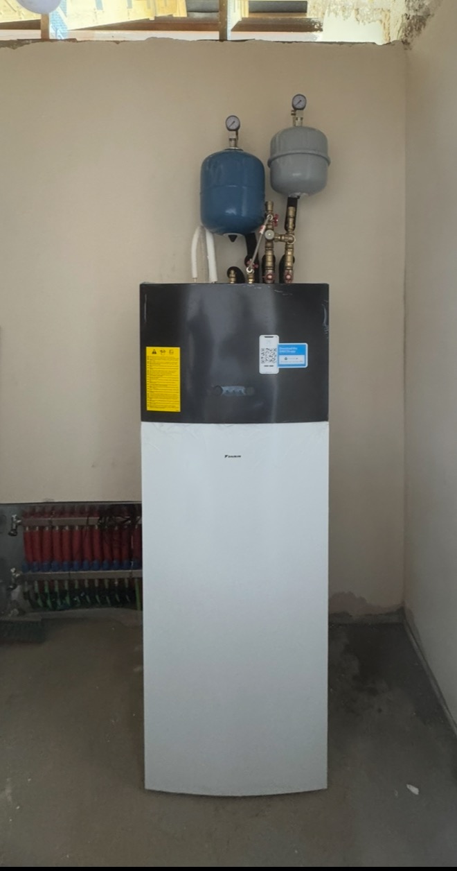 Biało-czarna pompa ciepła Daikin z dwoma zbiornikami wyrównawczymi, instalacja w surowym pomieszczeniu z widoczną rozdzielnią ogrzewania podłogowego.