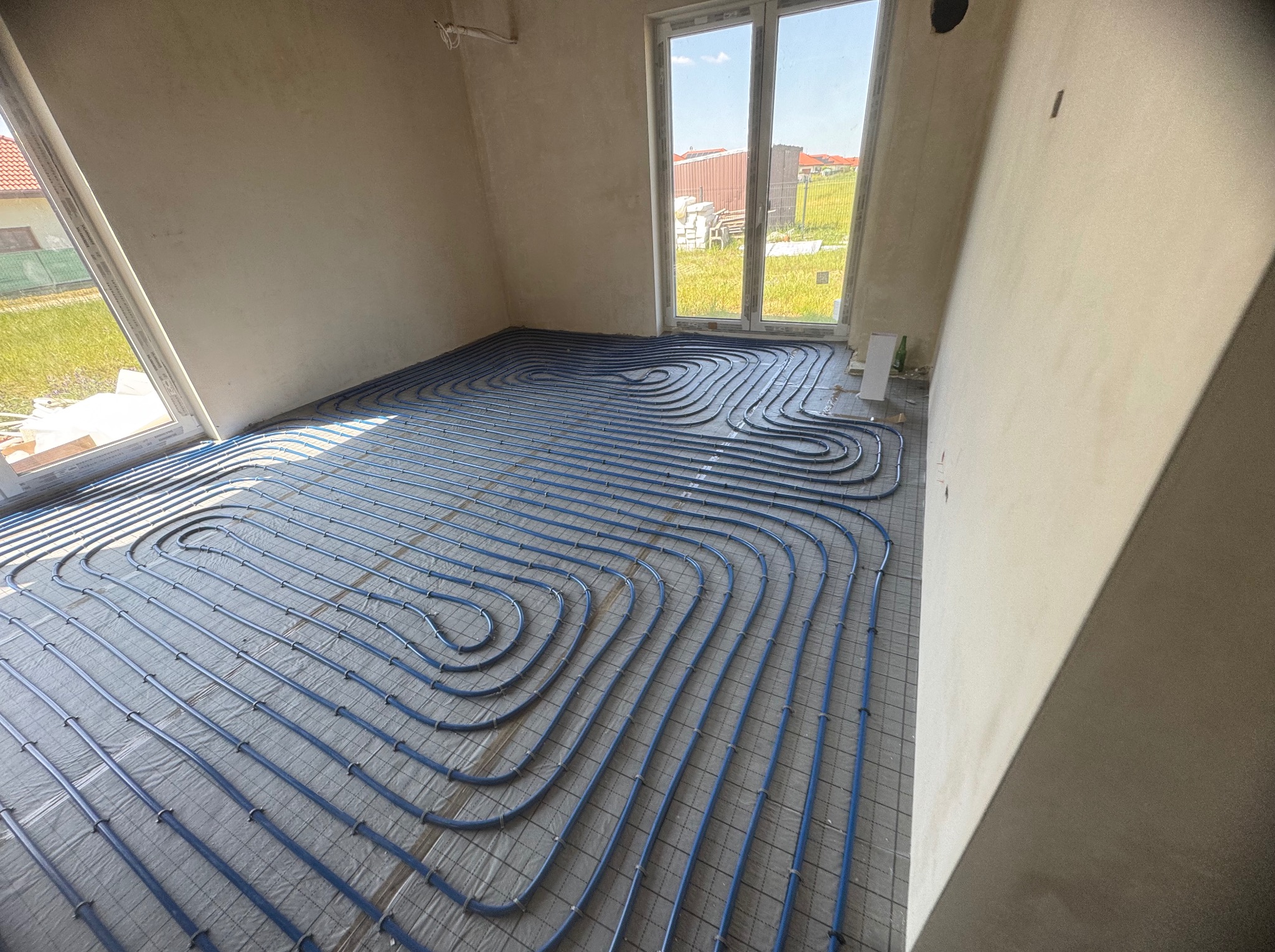 Niebieskie rury ogrzewania podłogowego ułożone na szarej macie w jasnym pomieszczeniu z oknami. Widok na zewnątrz z zielenią i domami.