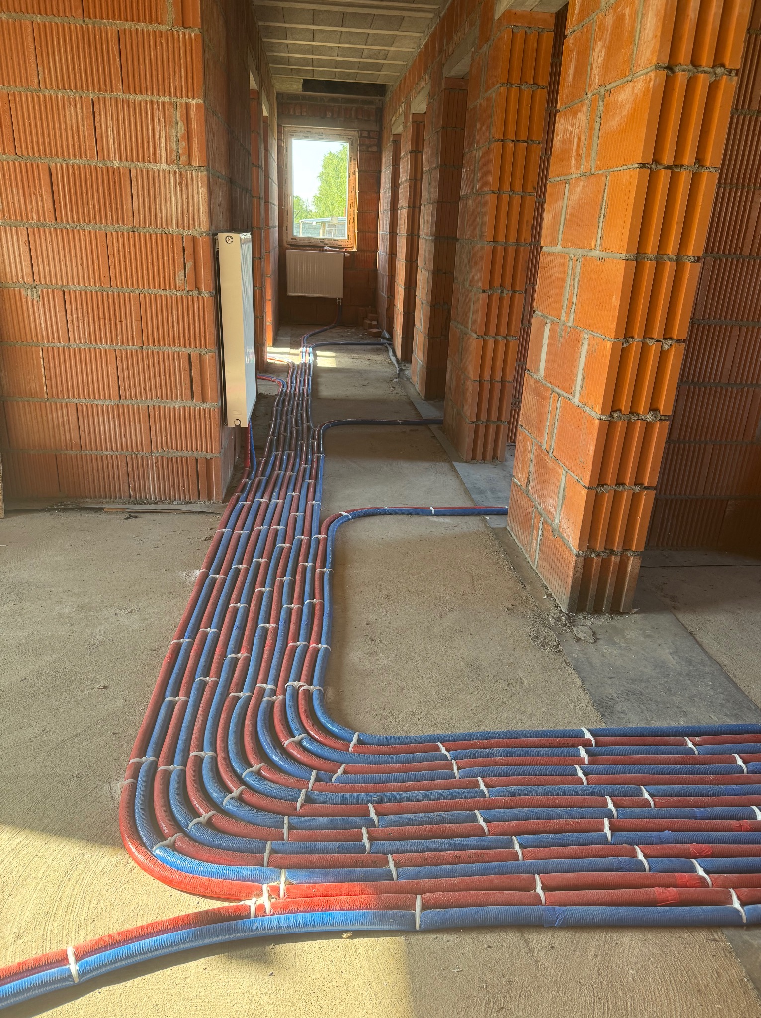Instalacja rur grzewczych (czerwono-niebieskich) w budynku w stanie surowym. Widoczne ceglane ściany i okno w tle. Rury ułożone równolegle, zabezpieczone białymi opaskami.