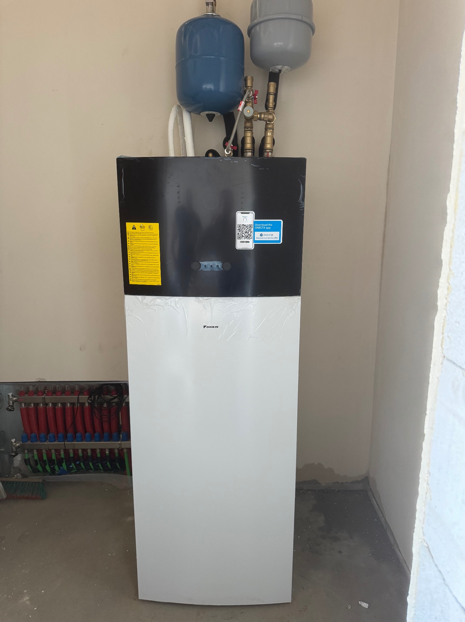 Nowoczesna pompa ciepła Daikin, z niebieskim i szarym zbiornikiem wyrównawczym. Widoczny rozdzielacz ogrzewania podłogowego, żółta naklejka ostrzegawcza i kod QR.