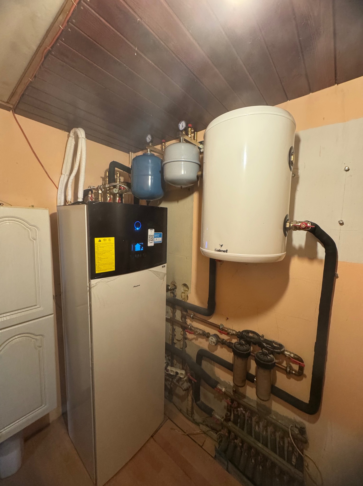 Biała pompa ciepła Daikin z zbiornikiem Galmet i instalacją hydrauliczną w pomieszczeniu z boazerią. Widoczne rury, filtry i manometry.