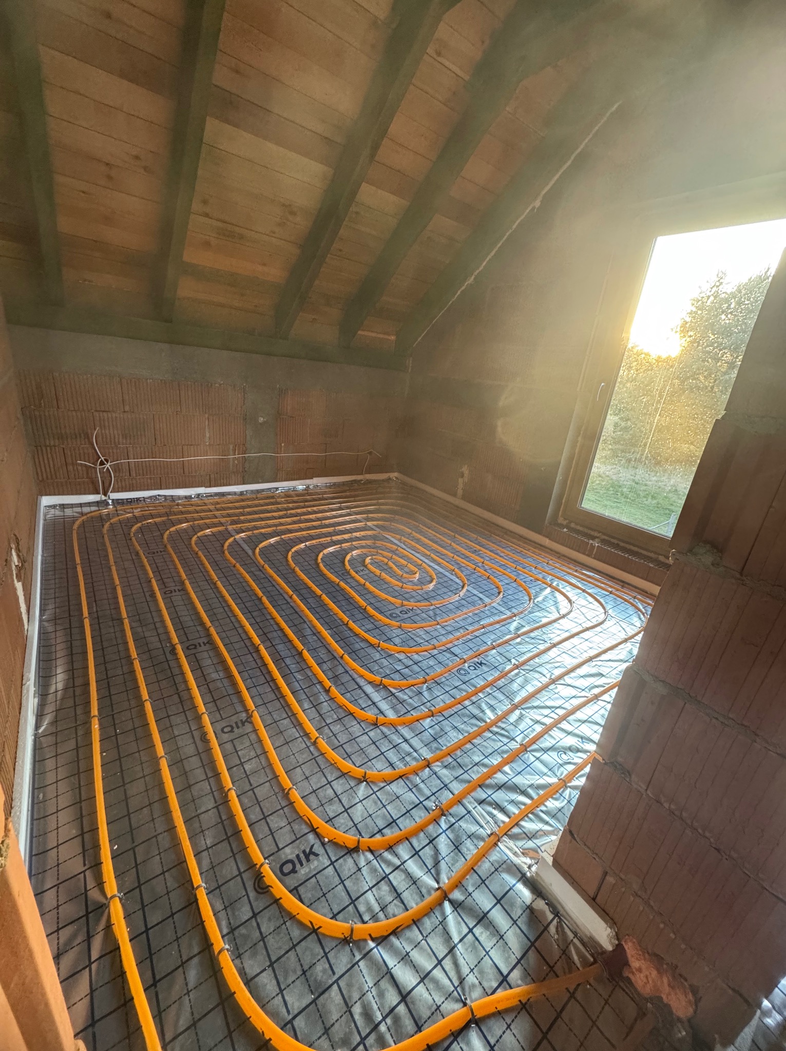 Ogrzewanie podłogowe w Oleśnicy: Pomarańczowe rury ułożone spiralnie na czarnej siatce w surowym wnętrzu poddasza, widok z góry, okno z promieniami słońca.