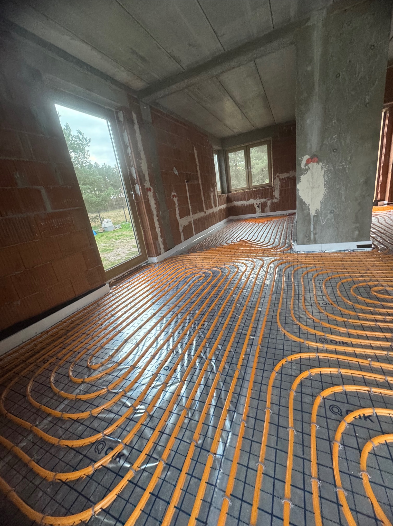Pomarańczowe rury ogrzewania podłogowego ułożone na szarej macie w surowym wnętrzu budynku z oknami. Widoczne cegły i betonowe elementy konstrukcyjne.