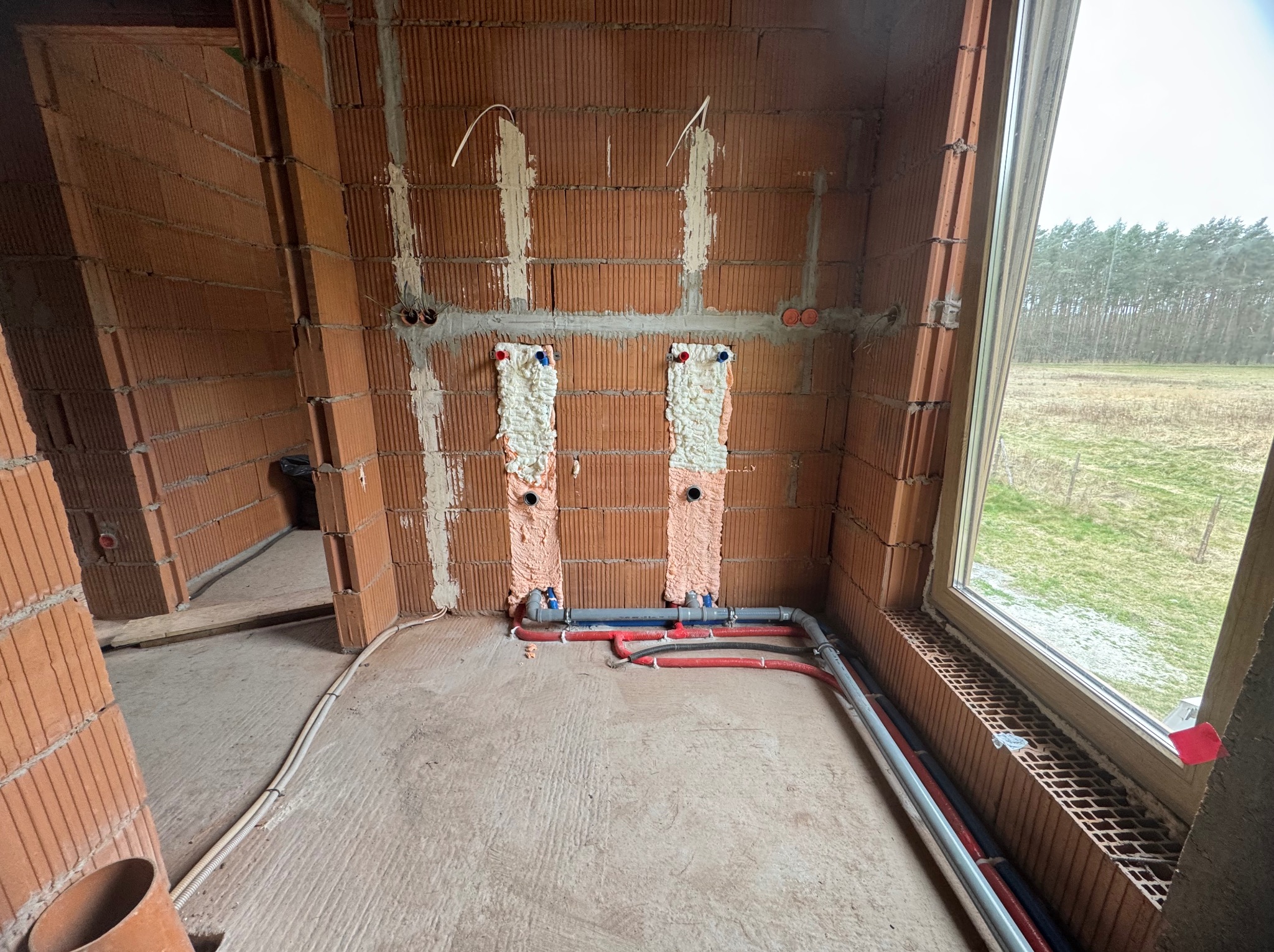 Montaż instalacji wodnej w surowym wnętrzu: czerwone i niebieskie rury przy ścianie z cegły, pianka montażowa wokół przyłączy. Widok na zewnątrz przez okno.