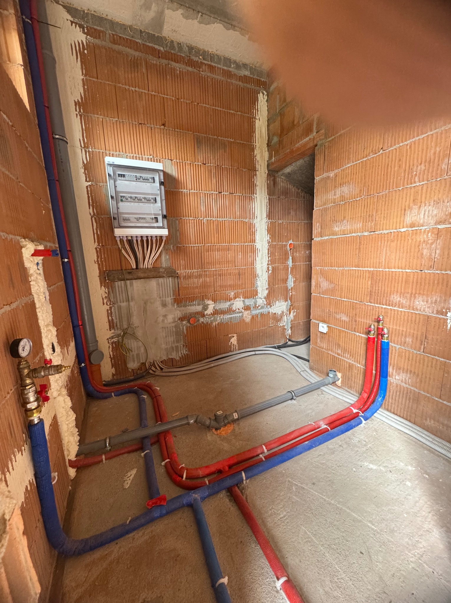 Instalacja wod-kan i elektryczna w surowym wnętrzu budynku z cegły. Widoczne rury w izolacji (czerwona i niebieska) oraz szara rura kanalizacyjna. Rozdzielnia elektryczna na ścianie.