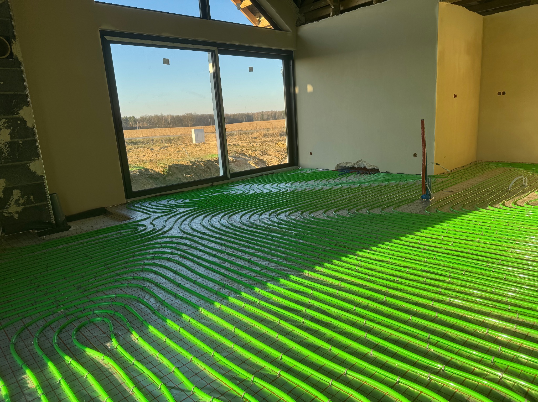 Zielone rury ogrzewania podłogowego ułożone na siatce w surowym wnętrzu z dużym oknem, przez które wpada światło słoneczne. Widoczna konstrukcja dachu.