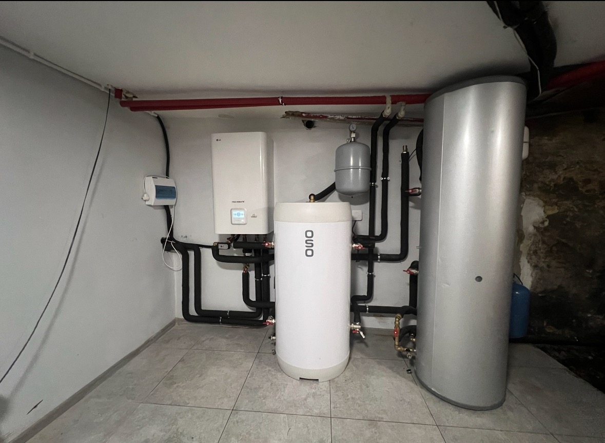 Zestaw urządzeń w kotłowni: biały piec grzewczy LG, biały zasobnik OSO, szary zbiornik wyrównawczy i srebrny bojler. Rury w izolacji, szara podłoga z płytek.