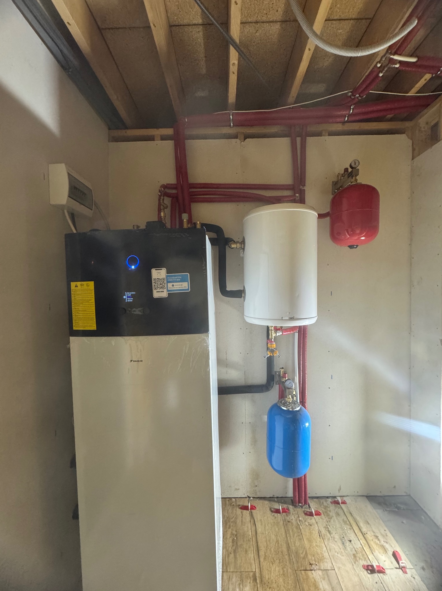 Instalacja pompy ciepła Daikin w kotłowni. Widoczne czerwone rury, zbiornik buforowy i elementy instalacji na drewnianej podłodze z elementami poziomującymi.