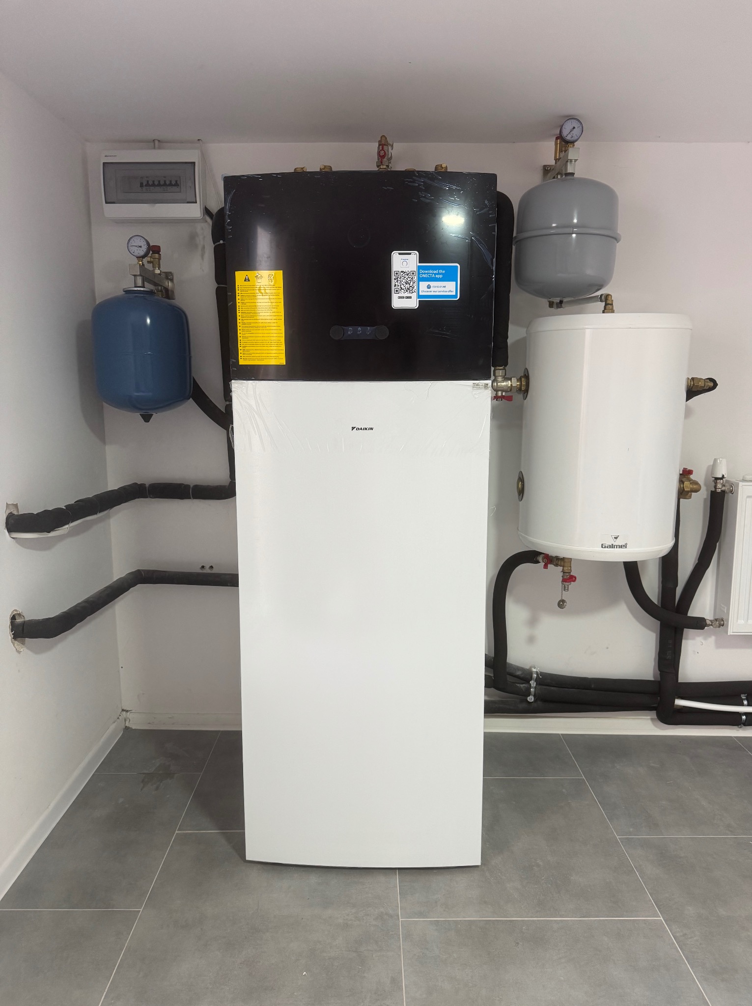 Zainstalowana pompa ciepła Daikin w pomieszczeniu technicznym z szarymi płytkami. Widoczne zbiorniki wyrównawcze i rury w izolacji. Tablica elektryczna na ścianie.