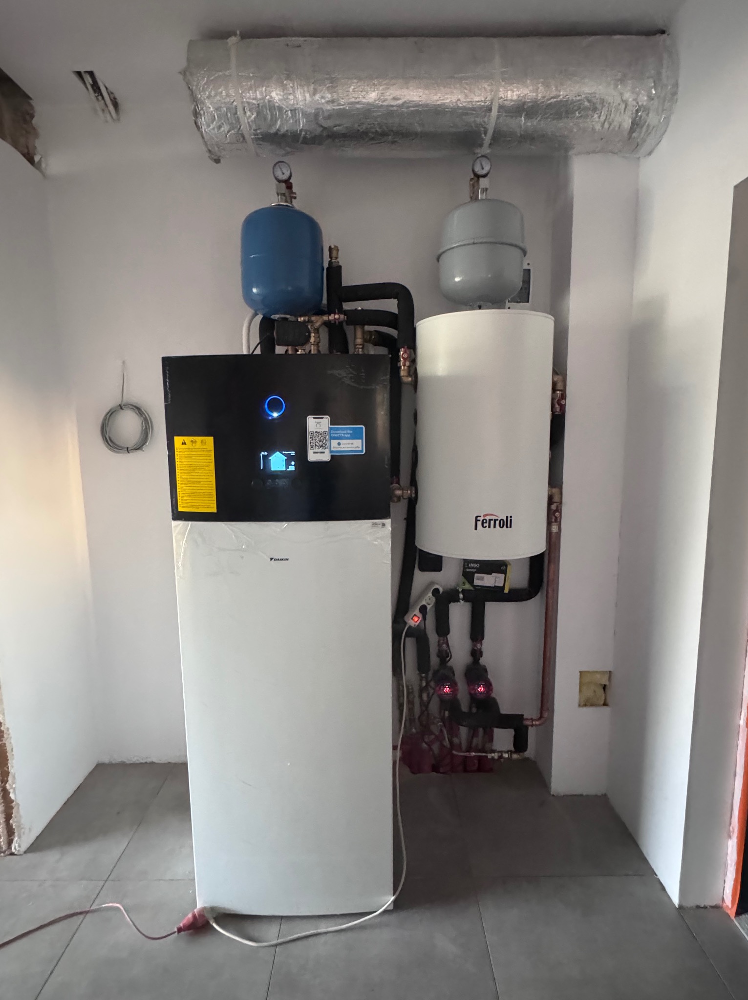 Nowoczesna instalacja grzewcza w kotłowni: pompa ciepła Daikin i zasobnik Ferroli. Widoczne rury, zbiorniki wyrównawcze i izolowany przewód wentylacyjny.