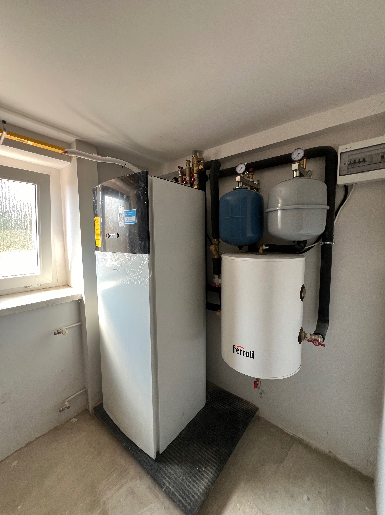 Biała pompa ciepła Ferroli, zbiorniki wyrównawcze i instalacja w pomieszczeniu z oknem. Część pompy owinięta folią ochronną, widoczne rury i zawory.