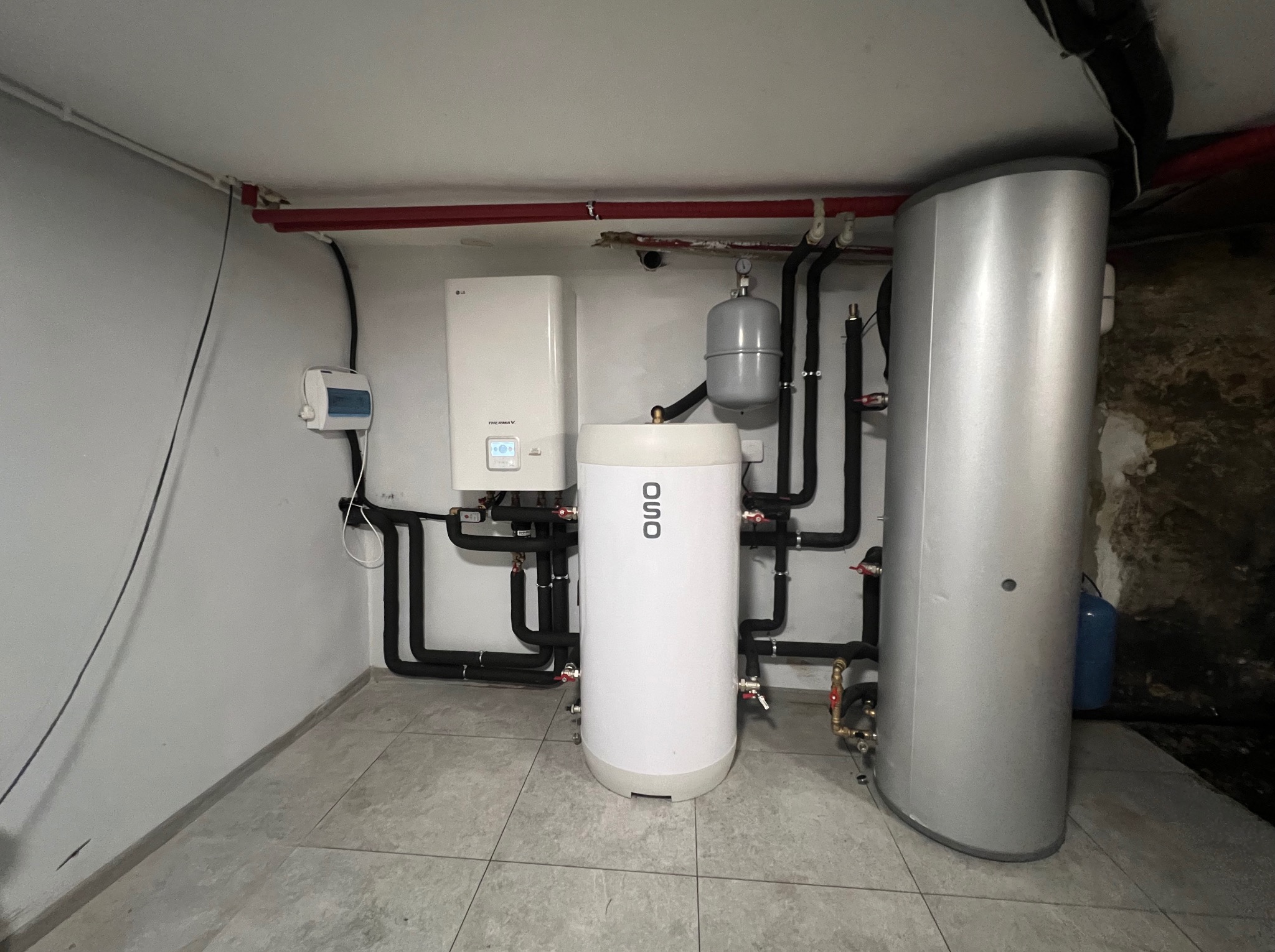 Zestaw urządzeń grzewczych w kotłowni: biały piec LG, biały zasobnik OSO i srebrny zbiornik, połączone czarnymi rurami. Widoczne instalacje na ścianach i suficie.