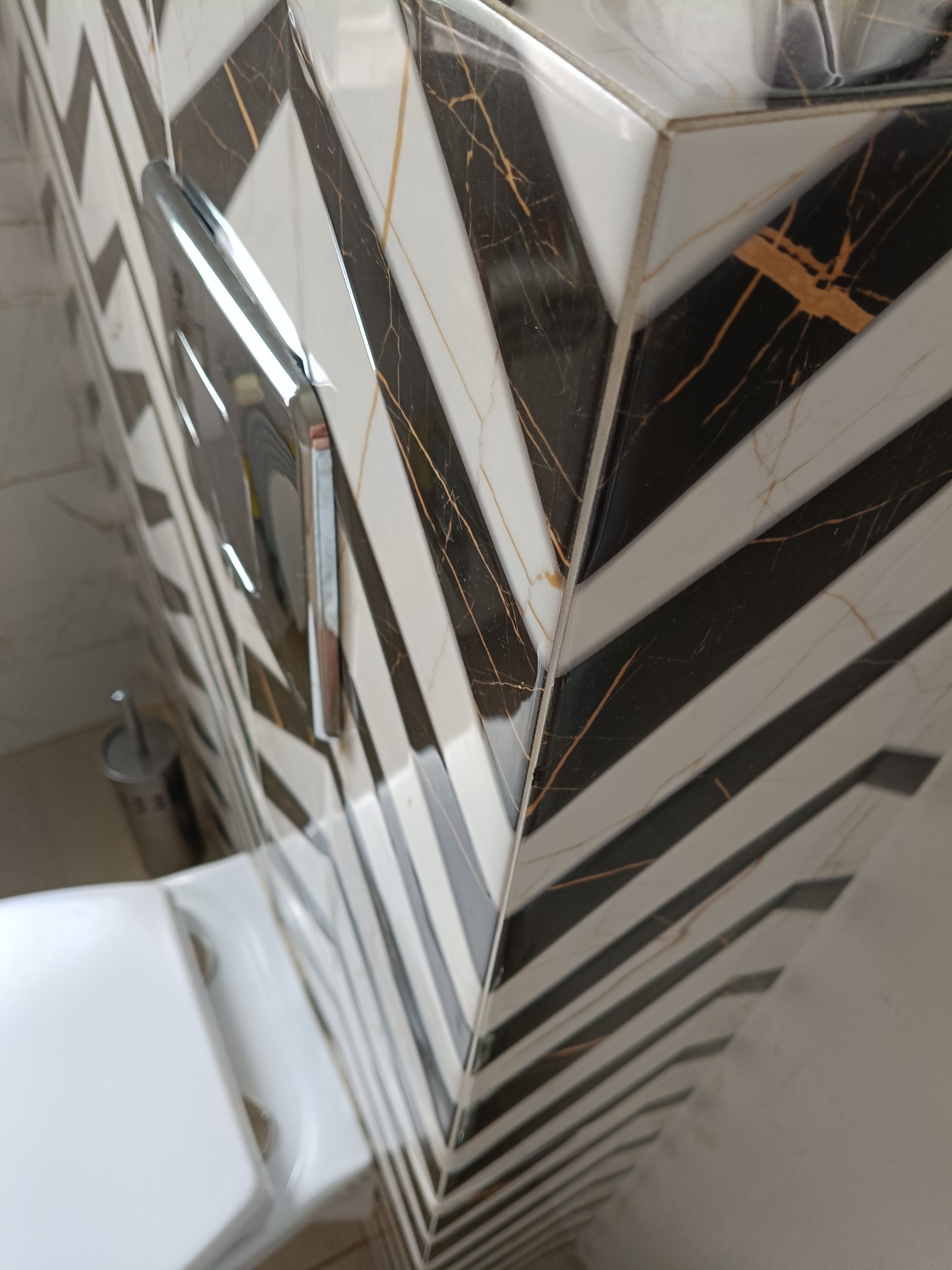 Wykończenie toalety z geometrycznym wzorem płytek w odcieniach bieli i czerni z subtelnymi złotymi żyłkami, widoczny fragment toalety i przycisk spłuczki.