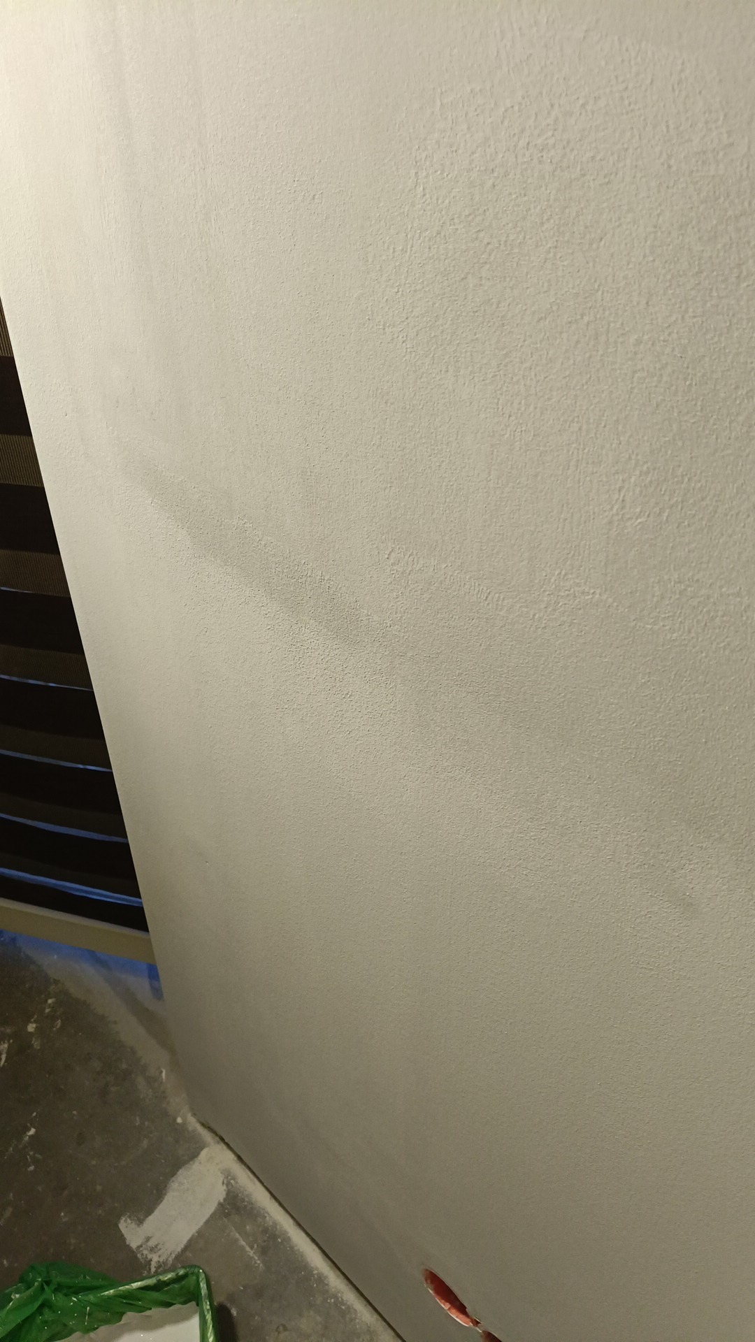 Ściana po malowaniu na jasny odcień, widoczna faktura. W tle żaluzje, na podłodze ślady farby. Gniazdko elektryczne w dolnym rogu kadru.