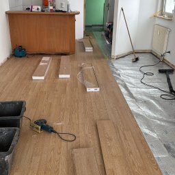 Wykończenia ,szpachlowanie ,malowanie ,płytki - Układanie paneli podłogowych w mieszkaniu, widoczne panele, narzędzia i częściowo ułożona podłoga. W tle aneks kuchenny i schody.