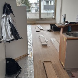 Wykończenia ,szpachlowanie ,malowanie ,płytki - Układanie jasnych paneli podłogowych w pokoju z widokiem na budynek. Częściowo ułożona podłoga, widoczne materiały i narzędzia. Prace w toku.