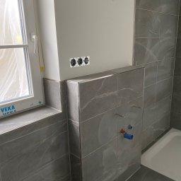 Wykończenia ,szpachlowanie ,malowanie ,płytki - Wykończona łazienka z szarymi płytkami imitującymi marmur, widoczne przyłącza wodne i gniazdka elektryczne. Okno z folią ochronną w rogu.