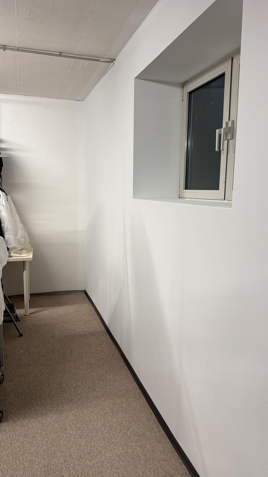 Wnętrze po odświeżeniu: białe ściany z oknem w grubym murze, brązowa wykładzina, czarna listwa przypodłogowa. Minimalistyczny wystrój, neutralna kolorystyka.