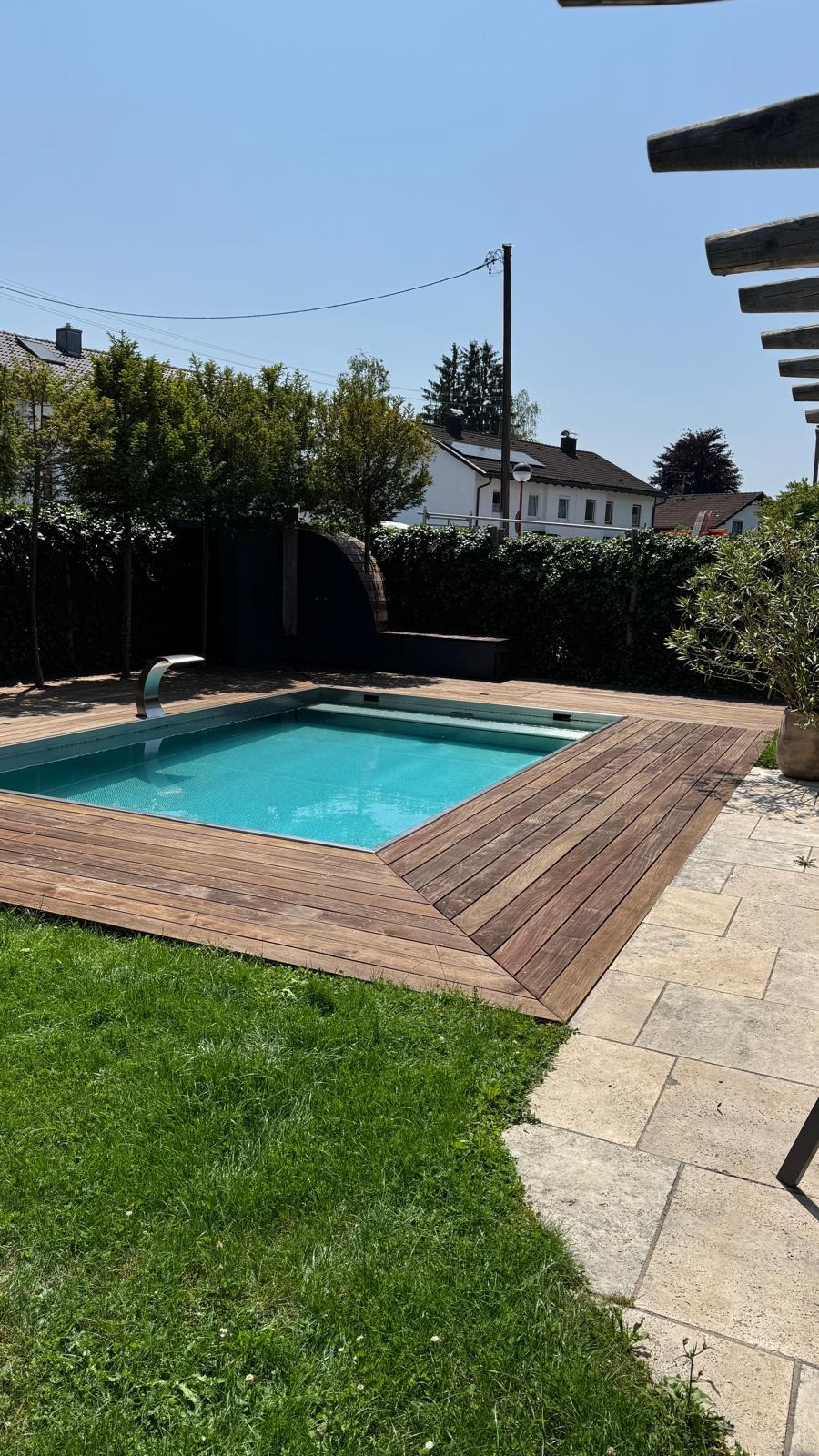 Nowoczesny basen ogrodowy z drewnianym tarasem i kamienną ścieżką w słoneczny dzień. Widoczne zielone trawniki i żywopłot w tle.