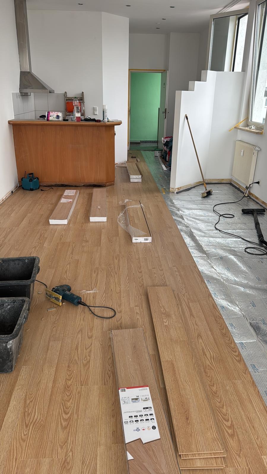 Układanie paneli podłogowych w mieszkaniu, widoczne panele, narzędzia i częściowo ułożona podłoga. W tle aneks kuchenny i schody.