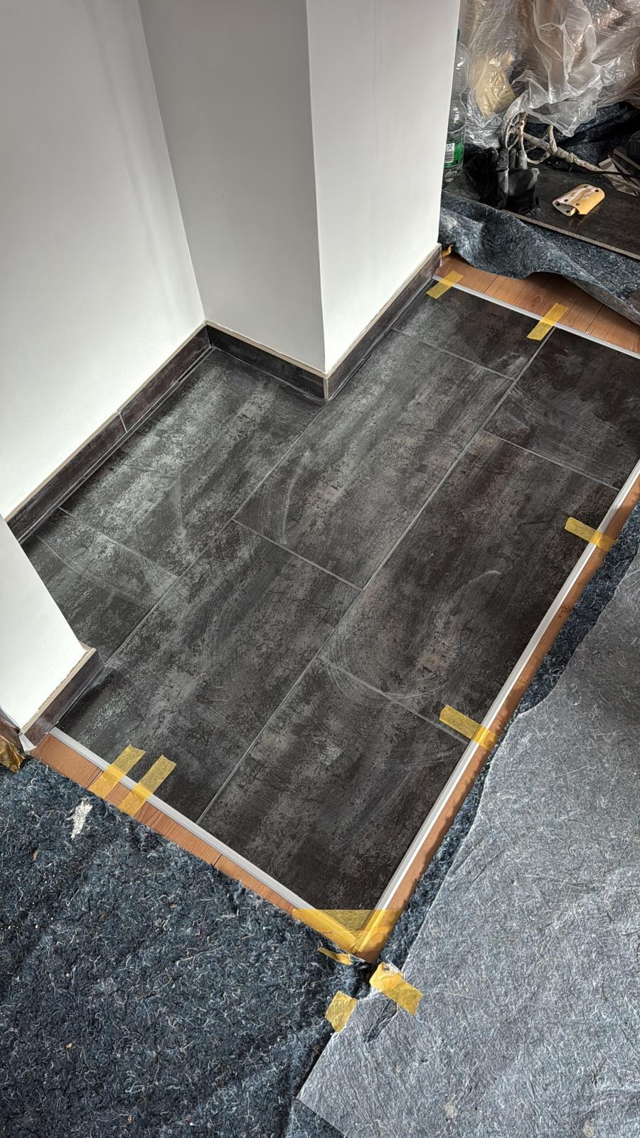 Fragment podłogi z ciemnymi płytkami w trakcie układania, zabezpieczona taśmą i folią ochronną, widoczne narzędzia i materiały budowlane.