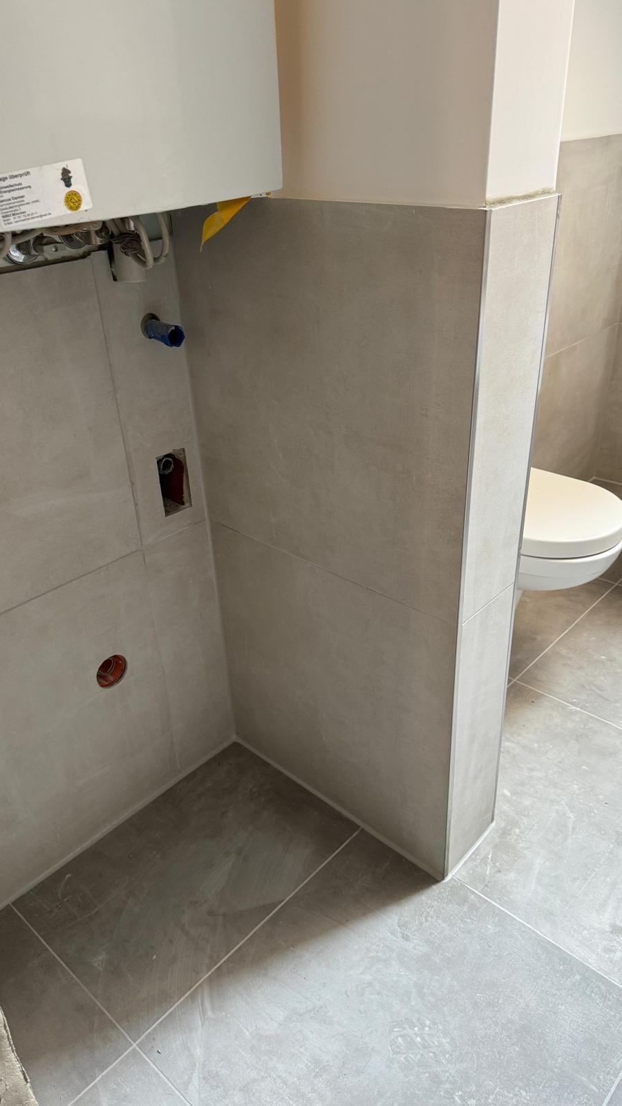 Wykończona łazienka z szarymi płytkami, widoczne narożniki wykończeniowe, fragment toalety, instalacja wodna i otwory pod gniazdka. Ujęcie z dołu, na pierwszym planie podłoga.