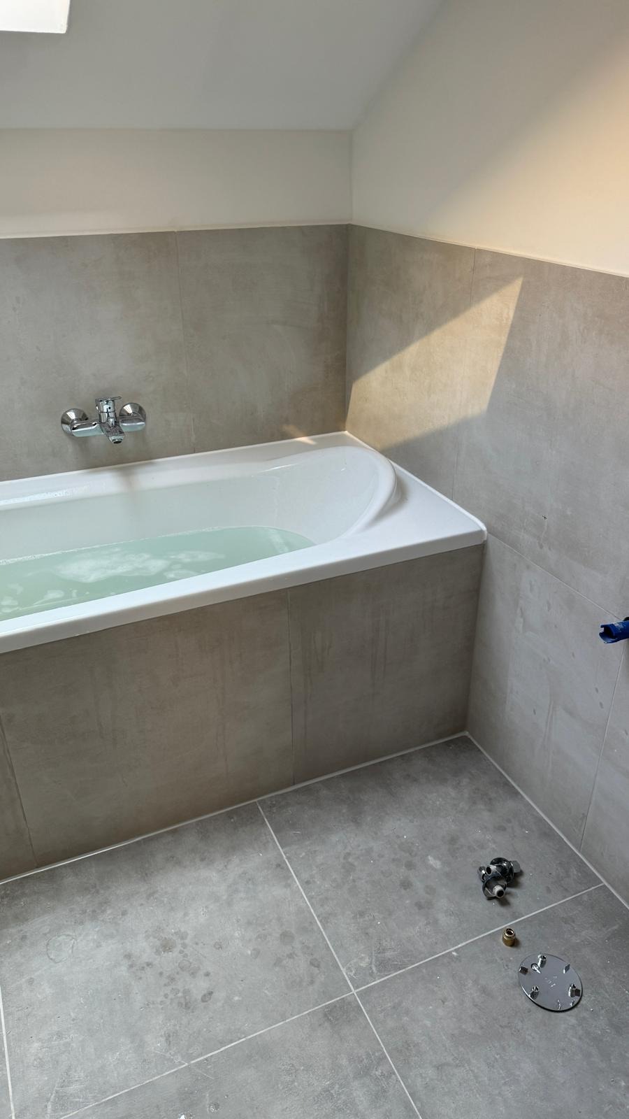 Wykończona łazienka z wanną wypełnioną wodą. Ściany i obudowa wanny wyłożone szarymi płytkami. Na podłodze widoczne elementy instalacyjne i plamy po zaprawie.