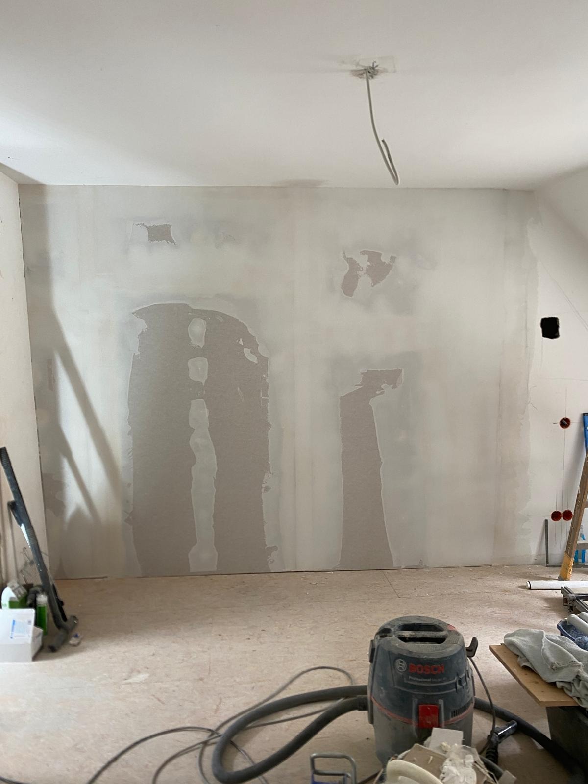 Ściana po szpachlowaniu z widocznymi łatami, przygotowana do malowania. Na podłodze widoczny odkurzacz przemysłowy i narzędzia.