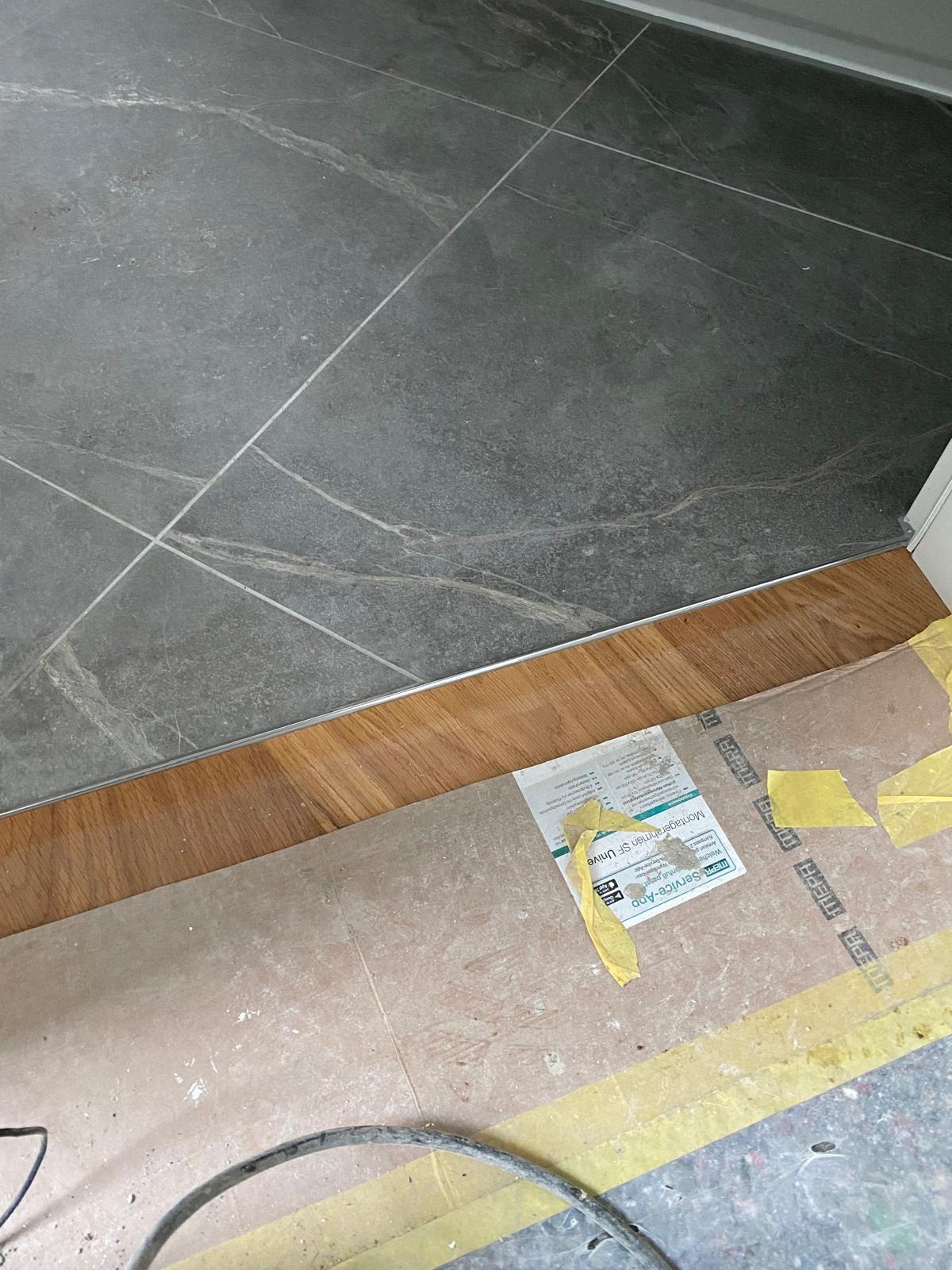 Położone szare płytki imitujące marmur, z widocznymi fugami, łączą się z drewnianą podłogą za pomocą srebrnej listwy. Częściowo widoczne zabezpieczenia podłogi.