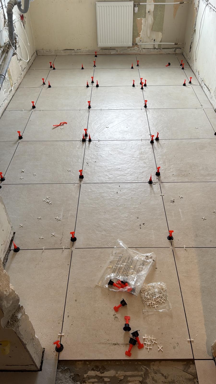 Układanie jasnych płytek podłogowych z użyciem systemu poziomowania, widoczne krzyżyki i kliny. Prace glazurnicze w trakcie realizacji w Gniezno.