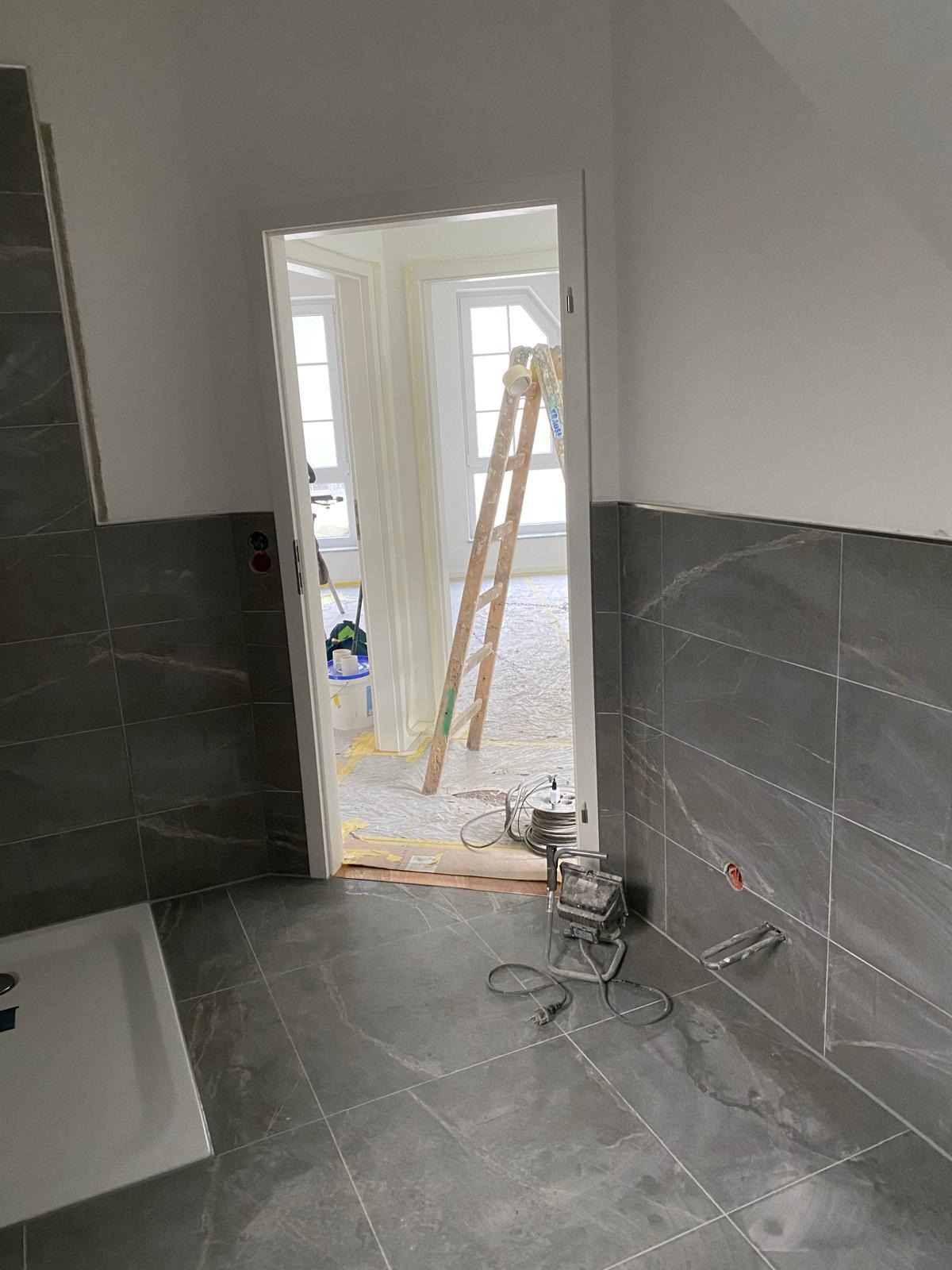 Wykończona łazienka z szarymi płytkami imitującymi marmur. Widoczna drabina malarska i narzędzia, sugerujące prace wykończeniowe. W tle okno z białymi ramami.