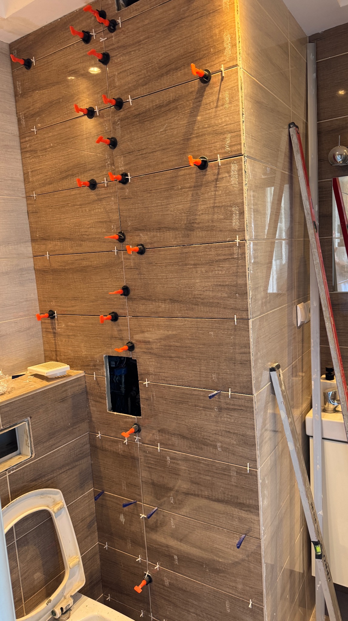 Proces układania brązowych płytek imitujących drewno w łazience, z widocznymi systemami poziomowania płytek i wyciętym otworem na instalację.