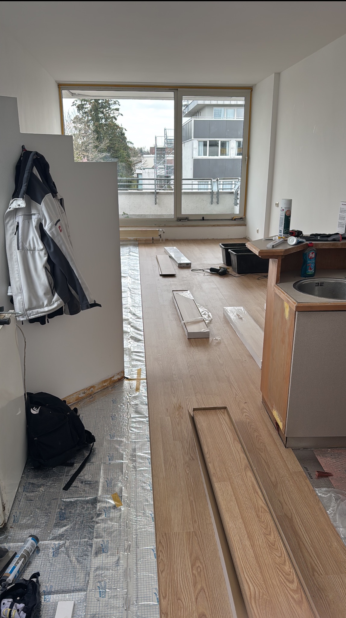 Układanie jasnych paneli podłogowych w pokoju z widokiem na budynek. Częściowo ułożona podłoga, widoczne materiały i narzędzia. Prace w toku.