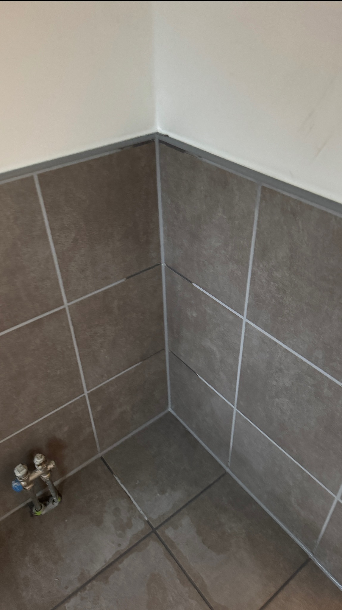 Wykończony narożnik łazienki z brązowymi płytkami ceramicznymi, fugami i fragmentem armatury. Widoczny styk płytek ze ścianą.