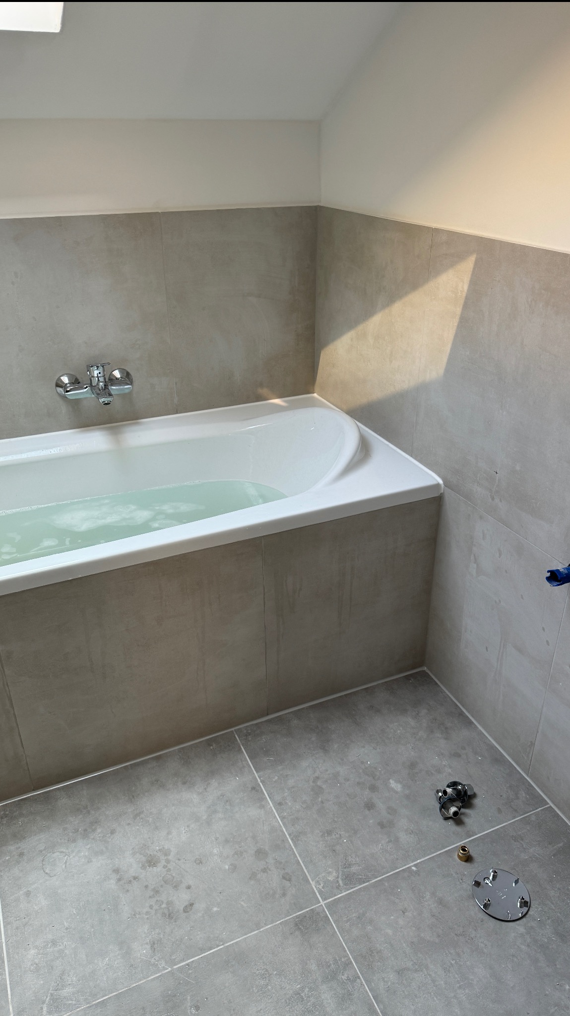 Wanna z wodą w łazience wyłożonej szarymi płytkami. Na podłodze widoczne elementy armatury. Świeże, jasne wnętrze po remoncie, z delikatnym cieniem na ścianie.