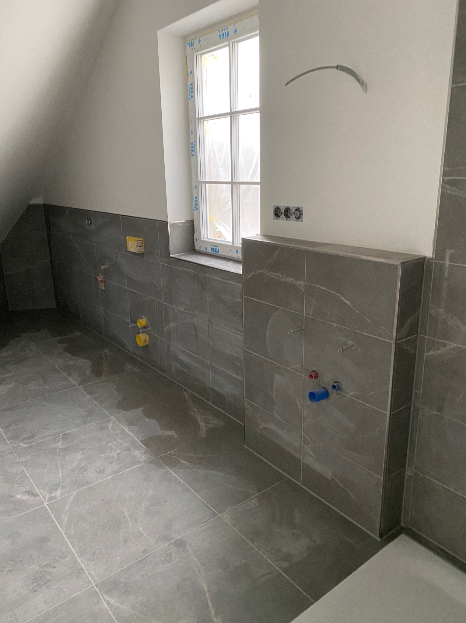 Wykończona łazienka na poddaszu: szare płytki imitujące marmur na podłodze i ścianach, widoczne przyłącza wodno-kanalizacyjne, okno dachowe z białą ramą.